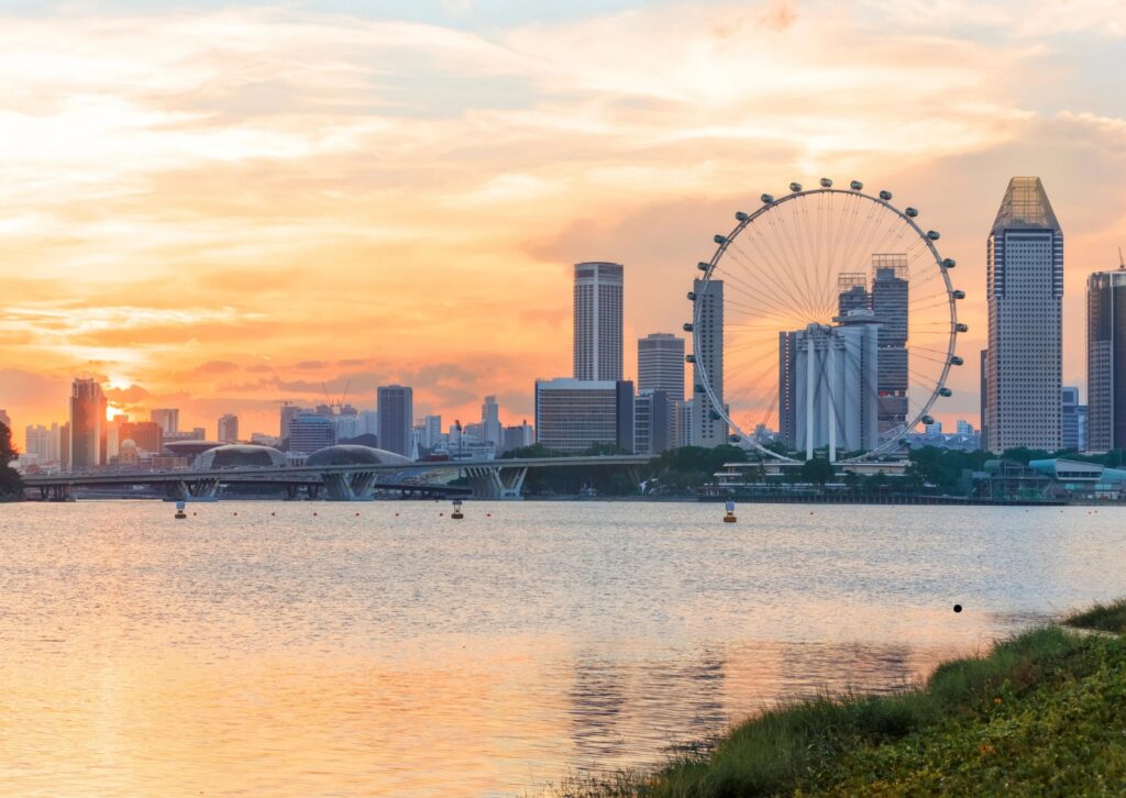 Singapore skyline at sunset with a prominent Ferris wheel in the foreground, showcasing vibrant colors and city lights.