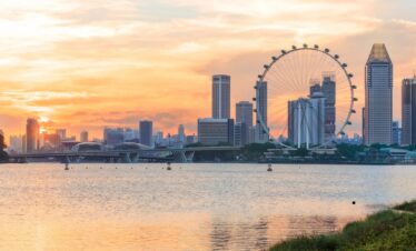 Singapore skyline at sunset with a prominent Ferris wheel in the foreground, showcasing vibrant colors and city lights.
