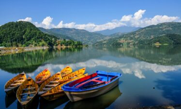 Three boats are moored on the edge of a tranquil lake, with trees lining the shore.