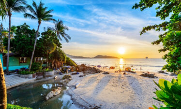 A serene beach in Thailand at sunset, with vibrant orange and pink hues reflecting on the calm water.